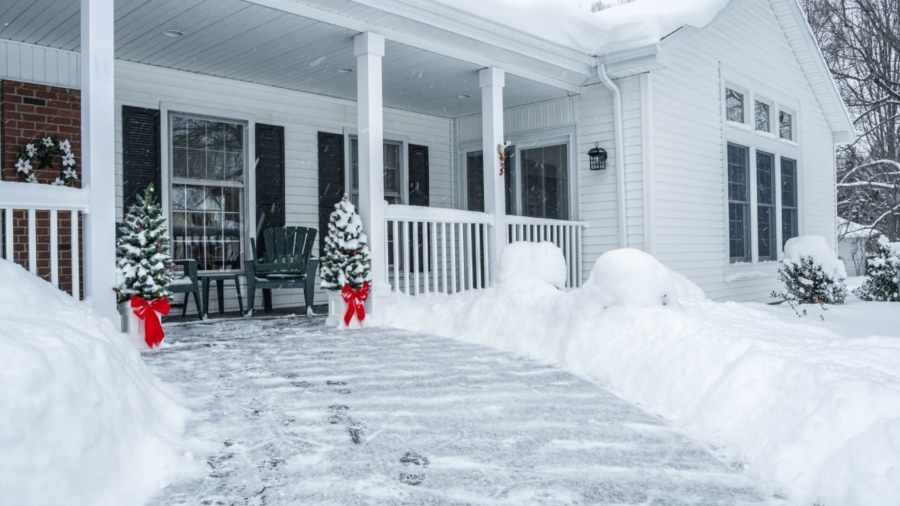 Deep snow has been shoveled, cleared and piled along the sides of the front yard footpath of this colonial style home. Two small holiday Christmas trees at the end of the walkway are decorated with Christmas ornaments and wide, bright red ribbon Christmas bows. Heavy blizzard snow continues to come down hard and blow around in all directions - already starting to re-coat the slippery surface and cover the fresh footprints. Nothing surprising here - just another early January winter snow storm in a rural suburban residential district neighborhood near Rochester, New York.