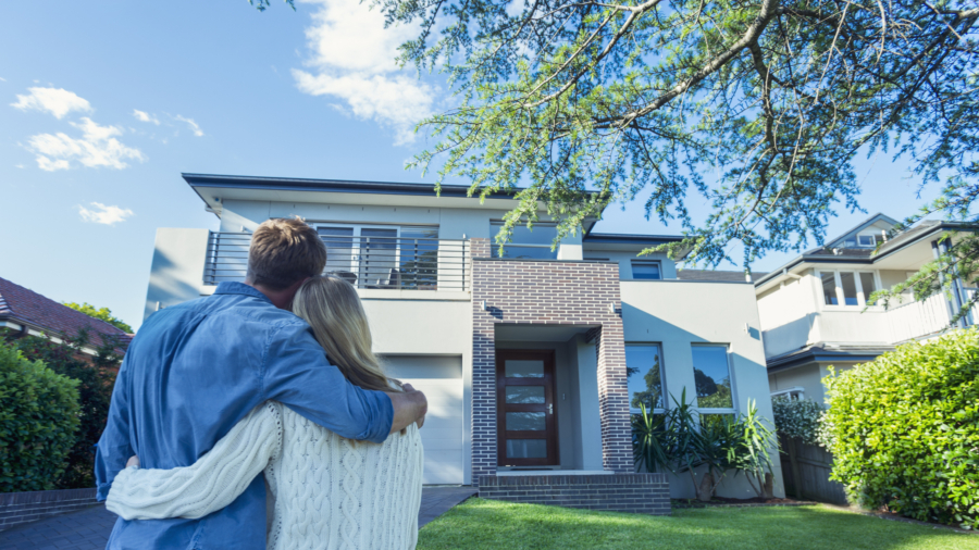 Couple standing in front of their new home. They are both wearing casual clothes and embracing. Rear view from behind them. The house is contemporary with a brick facade, driveway, balcony and a green lawn. The front door is also visible. Copy space