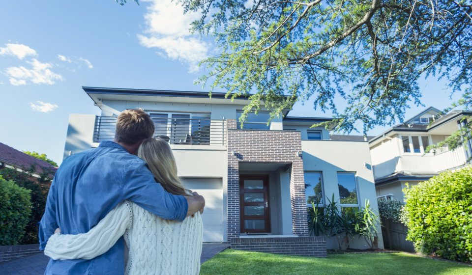 Couple standing in front of their new home. They are both wearing casual clothes and embracing. Rear view from behind them. The house is contemporary with a brick facade, driveway, balcony and a green lawn. The front door is also visible. Copy space
